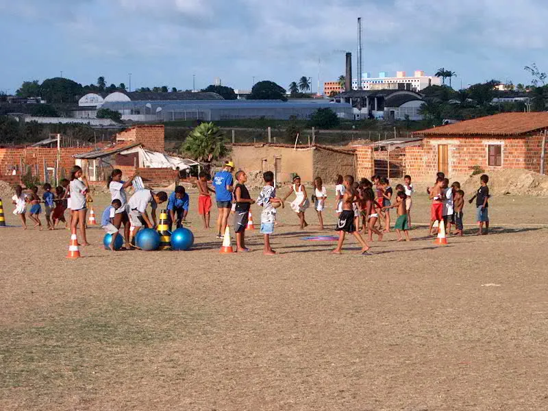 Kinderen en begeleiders in een sportactiviteit op een zandveld, met bakstenen huizen en de stad op de achtergrond.
